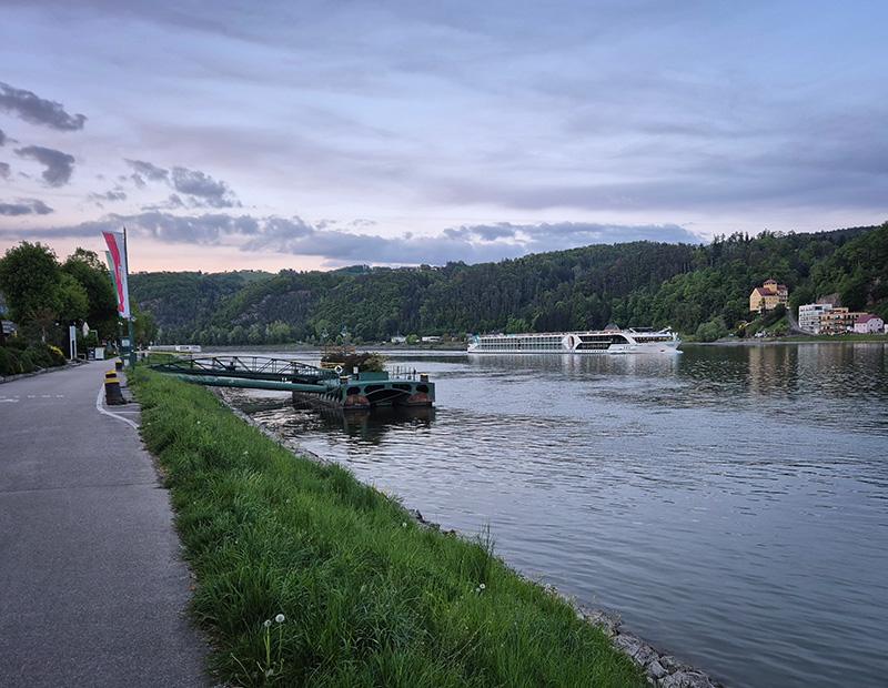 Kolesarska pot ob reki Donavi z rečno križarko in gozdnatimi pobočji ob mraku.